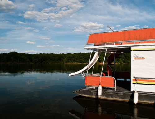A colorful houseboat docked on calm water, surrounded by green trees and a blue sky with scattered clouds.