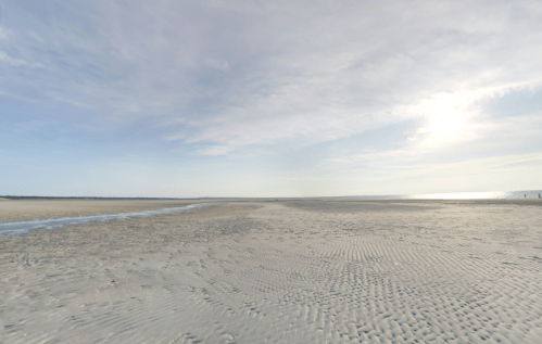 A serene beach scene with soft sand, gentle waves, and a clear sky, illuminated by the sun.
