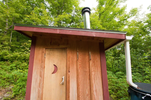 A wooden shed with a crescent moon door, surrounded by lush green trees, featuring a chimney on the roof.
