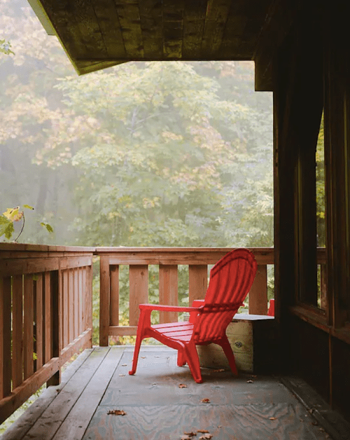 A red Adirondack chair on a wooden porch, surrounded by a misty forest with autumn foliage.