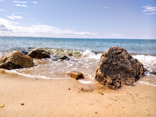 A serene beach scene with smooth rocks on the shore and gentle waves lapping at the sand under a clear blue sky.