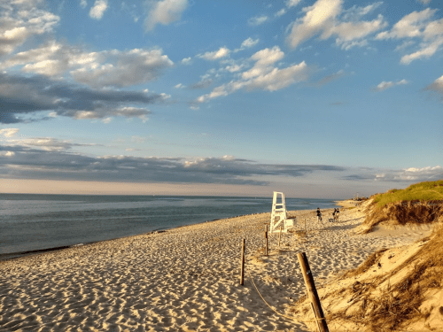 A serene beach scene with soft sand, gentle waves, and a lifeguard chair under a partly cloudy sky.