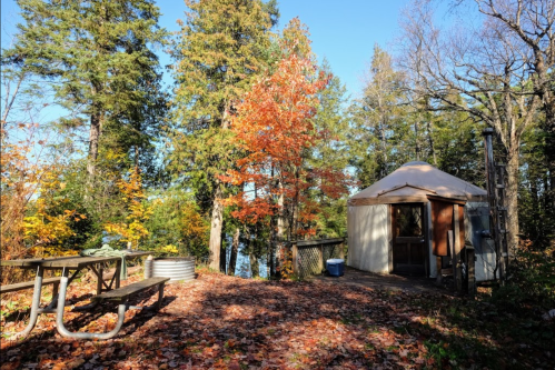A cozy yurt surrounded by colorful autumn trees and a picnic table near a serene lake.