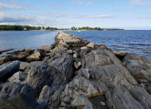 Rocky jetty extending into calm waters under a blue sky with scattered clouds.