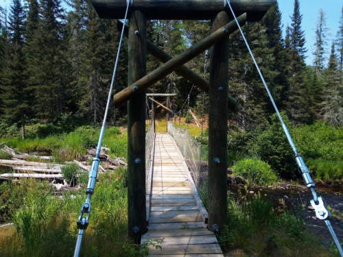 A wooden suspension bridge surrounded by trees and greenery, leading over a small stream.
