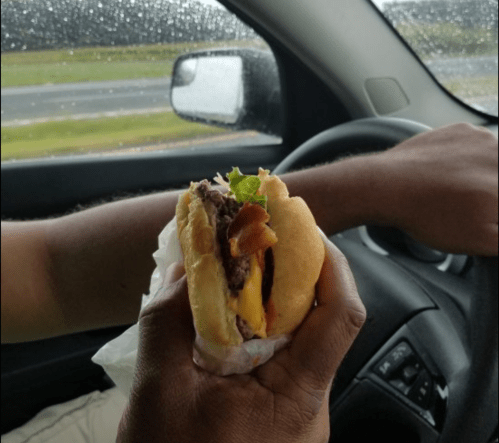 A hand holding a cheeseburger with bacon, while driving in a car on a rainy day.
