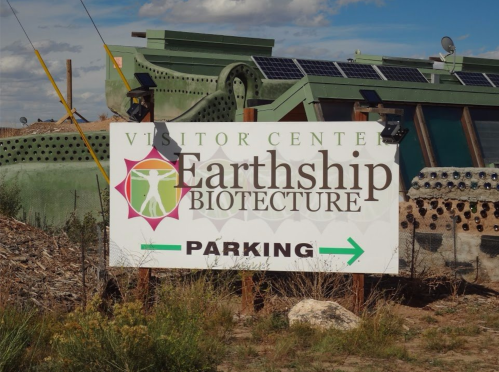Sign for Earthship Biotecture Visitor Center with a parking arrow, surrounded by natural landscape and solar panels.