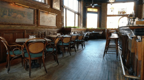 Cozy restaurant interior with wooden floors, empty tables, and large windows letting in natural light.