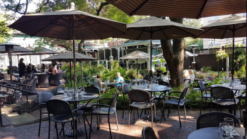 A cozy outdoor restaurant patio with tables, umbrellas, and lush greenery, filled with diners enjoying their meals.