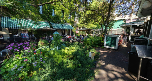 A vibrant outdoor dining area with green foliage, colorful flowers, and patrons enjoying their meals under umbrellas.
