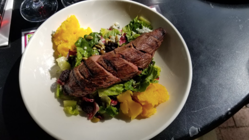 A plate featuring grilled steak on a bed of mixed greens, with yellow squash and garnished with seeds and dried fruits.