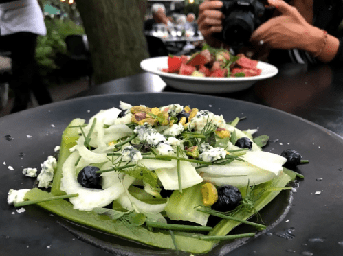 A fresh salad with celery, blue cheese, pistachios, and blueberries on a black plate, with another dish in the background.