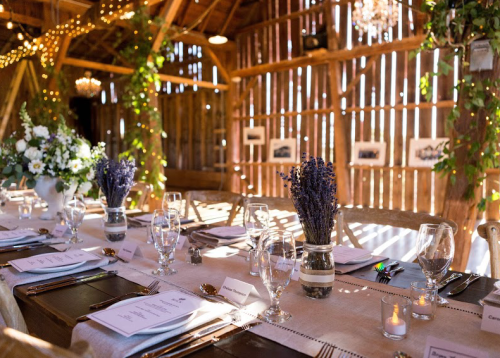 A beautifully decorated barn interior with a long dining table, flowers, candles, and rustic wooden walls.