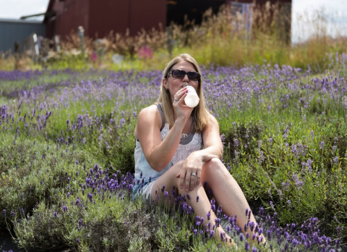 A woman sits in a lavender field, sipping a drink and enjoying the sunny day.