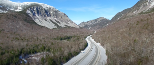 A snowy mountain landscape with a winding road surrounded by bare trees and towering peaks under a clear blue sky.