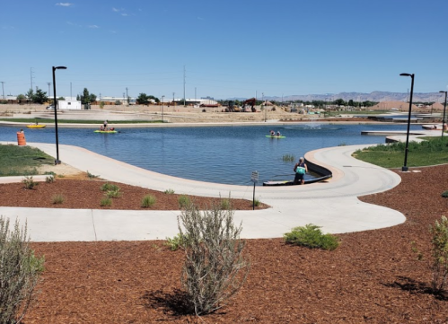 A serene park scene with a calm pond, walking paths, and people kayaking under a clear blue sky.