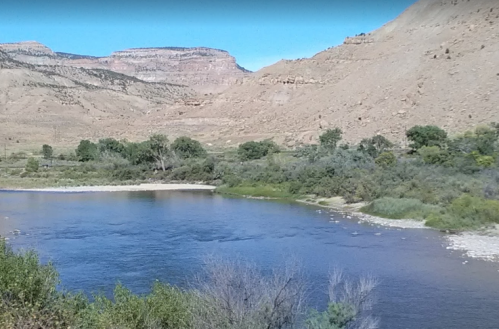 A serene river flows through a rocky landscape, surrounded by greenery and hills under a clear blue sky.