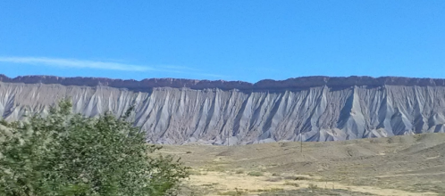 A rugged mountain range with steep, textured cliffs under a clear blue sky, framed by sparse vegetation in the foreground.