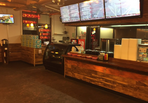 Interior of a casual restaurant with a wooden counter, menu screens, and a display case. Soft drinks and condiments are visible.