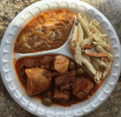 A paper plate with a serving of chicken stew, pasta salad, refried beans, and a slice of potato.