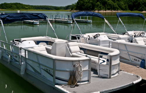 Two white pontoon boats docked at a calm lake, surrounded by green trees and a clear blue sky.