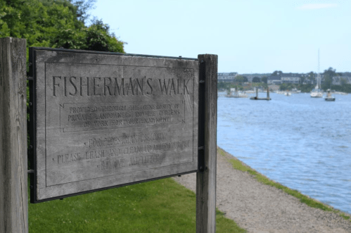 Sign for Fisherman's Walk by a river, with boats in the background and greenery along the shore.