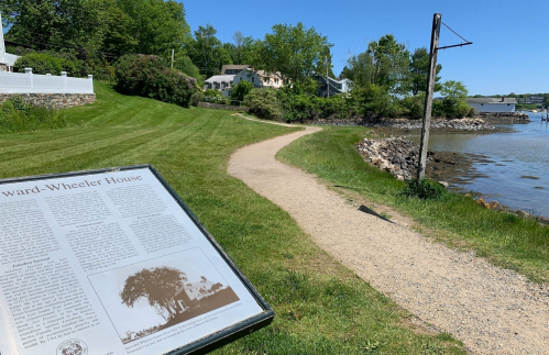 A grassy path leads to the water, with a historical sign about the Edward-Wheeler House in the foreground.