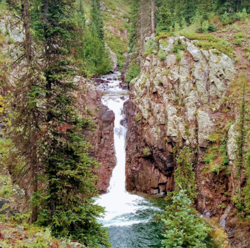 A waterfall cascades through a rocky gorge, surrounded by lush green trees and rugged terrain.
