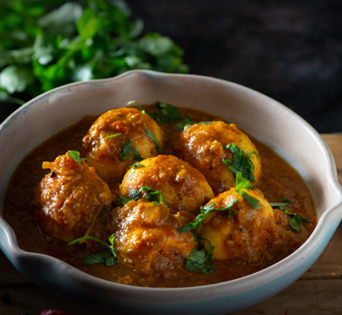 A bowl of spicy egg curry garnished with fresh cilantro, served on a wooden surface with herbs in the background.