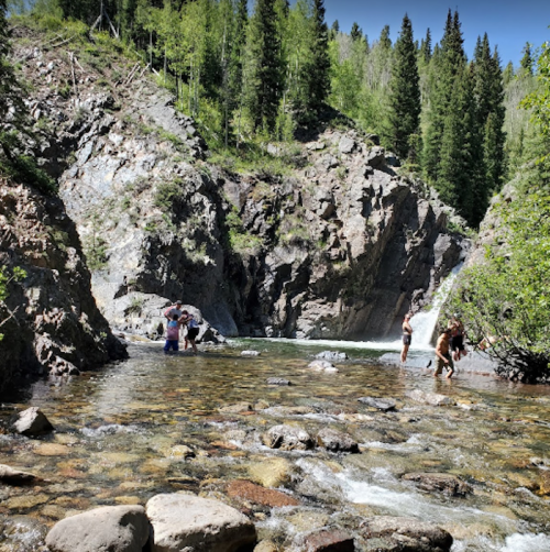 A serene mountain scene with people wading in a clear stream surrounded by rocky cliffs and lush green trees.
