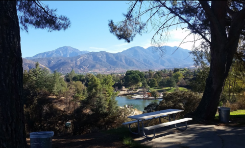 Scenic view of a lake surrounded by trees and mountains under a clear blue sky, with a picnic table in the foreground.