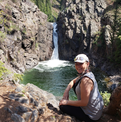 A woman sits on a rock by a waterfall, surrounded by lush greenery and rocky cliffs.
