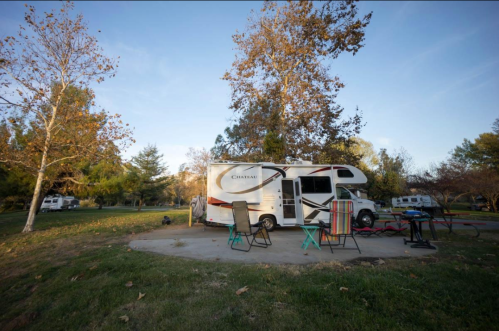 A parked RV in a grassy area with chairs and a table, surrounded by trees and a clear blue sky.