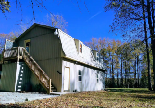 A modern two-story house with a metal roof, wooden stairs, and surrounded by trees under a clear blue sky.