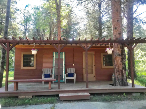A cozy wooden cabin surrounded by trees, featuring a porch with two chairs and hanging flower pots.