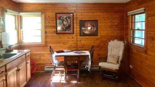 Cozy wooden cabin interior with a dining table, rocking chair, and framed artwork on the walls. Bright natural light.