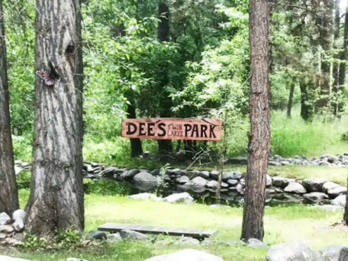 Wooden sign reading "Dees Twin Lakes Park" surrounded by trees and a small rocky stream.