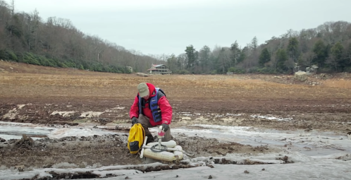 A person in a red jacket kneels on dry, cracked ground, preparing equipment near a low water level in a rural area.