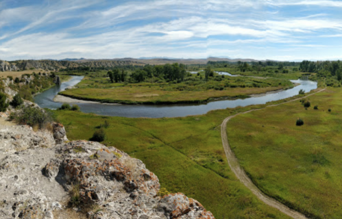 A panoramic view of a winding river surrounded by lush green fields and trees under a blue sky with scattered clouds.