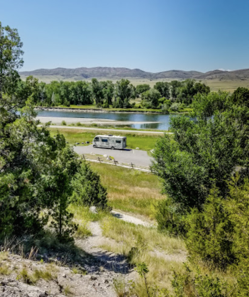 A scenic view of a river with an RV parked nearby, surrounded by lush greenery and distant mountains.