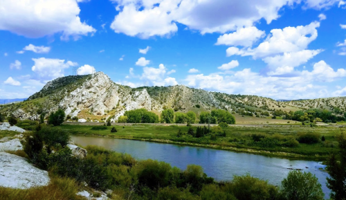 A serene river flows through a lush valley, framed by rocky hills and a bright blue sky with fluffy clouds.