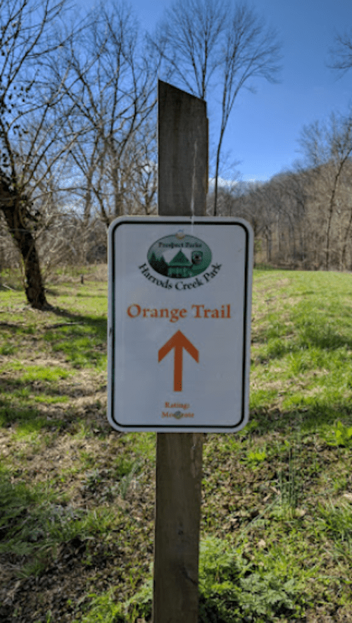 Sign for the Orange Trail at Harolds Creek Park, featuring an upward arrow and a scenic background of trees.