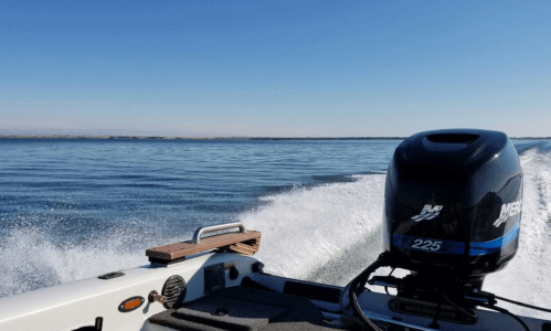 A boat's motor creates a wake on a calm lake under a clear blue sky.
