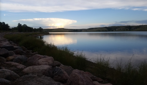 A serene lake scene with rocky shore, reflecting clouds and hills under a blue sky at sunset.