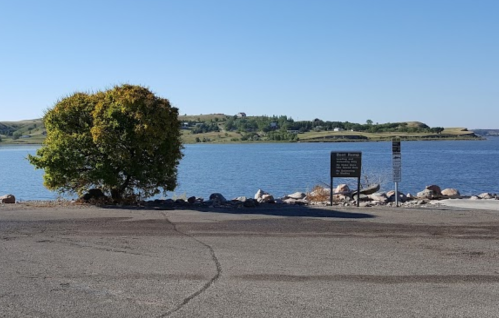 A serene lake view with a tree in the foreground and a sign near the water's edge, surrounded by green hills.