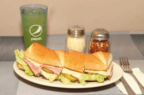 A sandwich with lettuce, ham, and cheese on a plate, accompanied by a green drink and condiments.