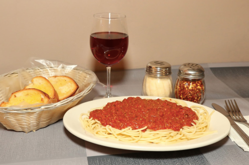 A plate of spaghetti with marinara sauce, a glass of red wine, and a basket of bread on a table.