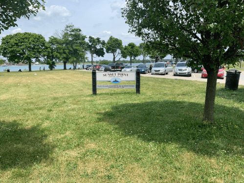 Sign for Sunset Point in a grassy area with trees and parked cars, near a waterfront.