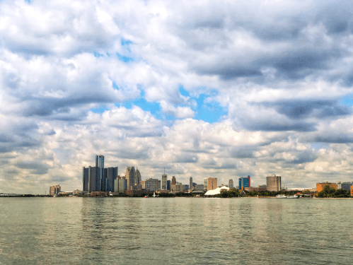 City skyline with tall buildings reflected in water under a cloudy sky.