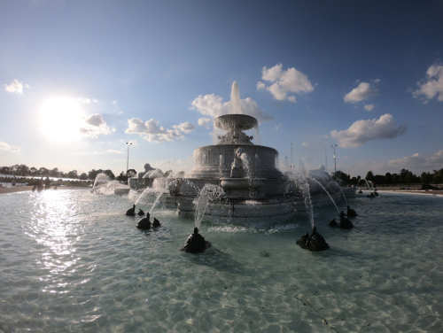 A large fountain with multiple tiers and water jets, surrounded by a clear pool, under a bright sky with clouds.
